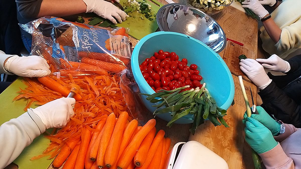 Ein Tisch mit frischem Gemüse, mehrere Hände, die das Gemüse zum kochen schneiden